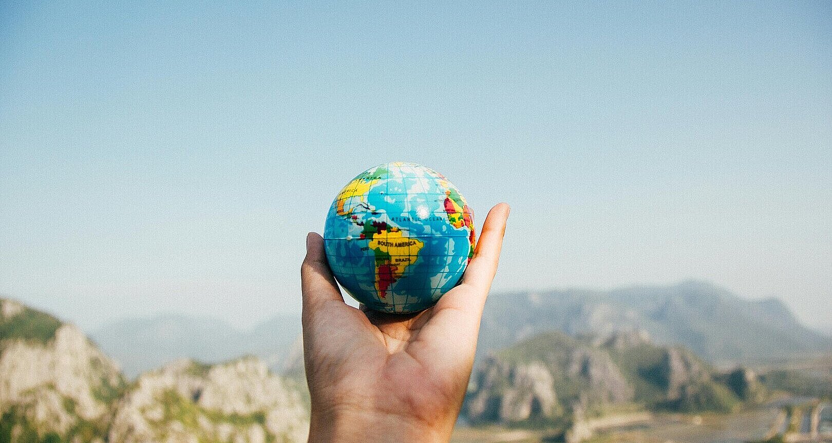 Hand hält einen kleinen Globus, im Hintergrund ein weites Tal