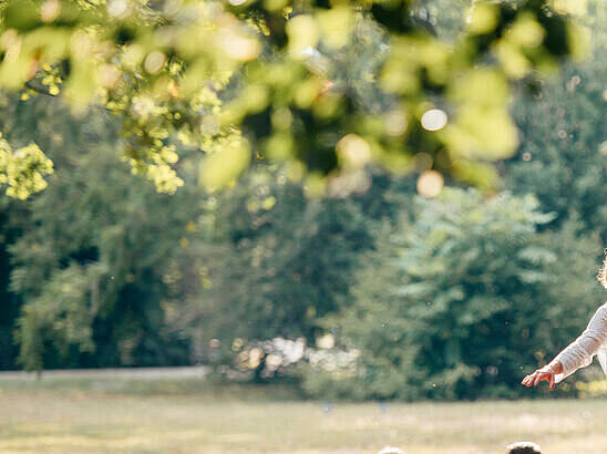 Eine junge Frau balanciert auf einer Slackline im Park, ein junger Mann hilft ihr